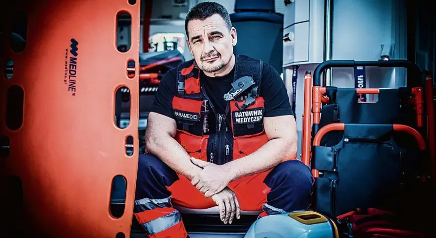 Ratownik medyczny w kamizelce z napisem "Paramedic" i "Ratownik Medyczny" siedzi w karetce obok noszy.