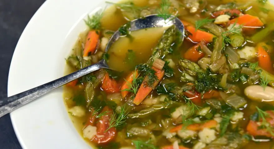 Un fumante minestrone di verdure, ricco di carote, fagioli e erbe aromatiche, servito in una ciotola bianca con un cucchiaio.