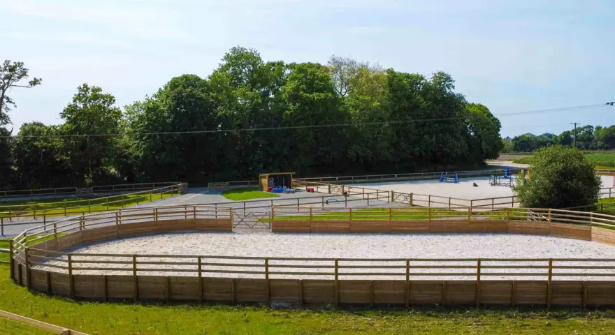 Un manège équestre, un rond d'Havrincourt, avec une piste en sable entourée de bois. Des obstacles d'équitation sont visibles au loin.