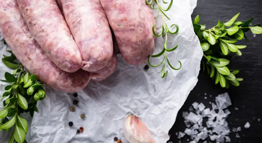 Surowa biała kiełbasa na papierze, obok gałązki rozmarynu i czosnek. Zastanawiasz się nad biała kiełbasa kcal?