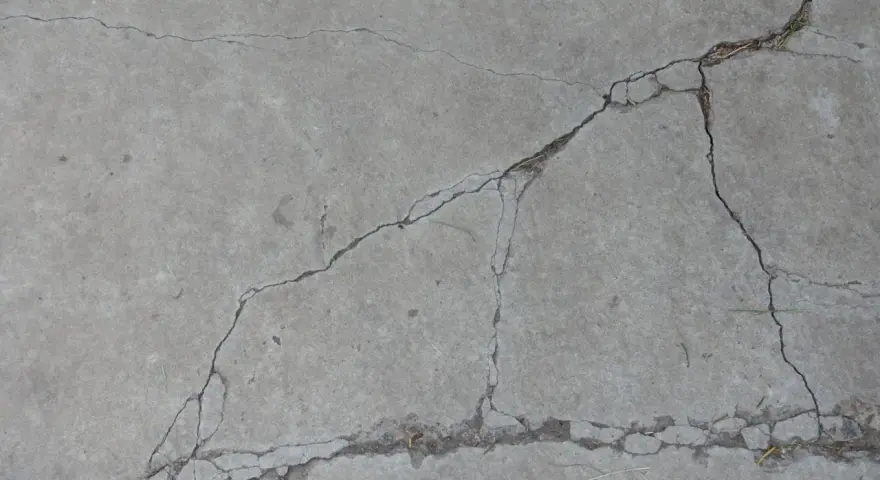 Popękany beton, jakby po długim okresie podlewania, z widocznymi szczelinami i drobnymi źdźbłami trawy.