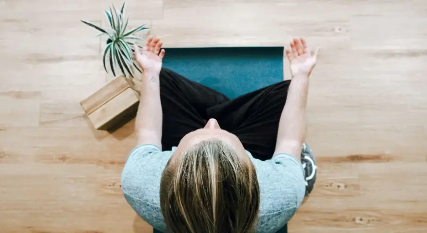 Person mediterar på en yogamatta, en grön växt och träblock bredvid. Perfekt för avkoppling på arbetsplatsen.