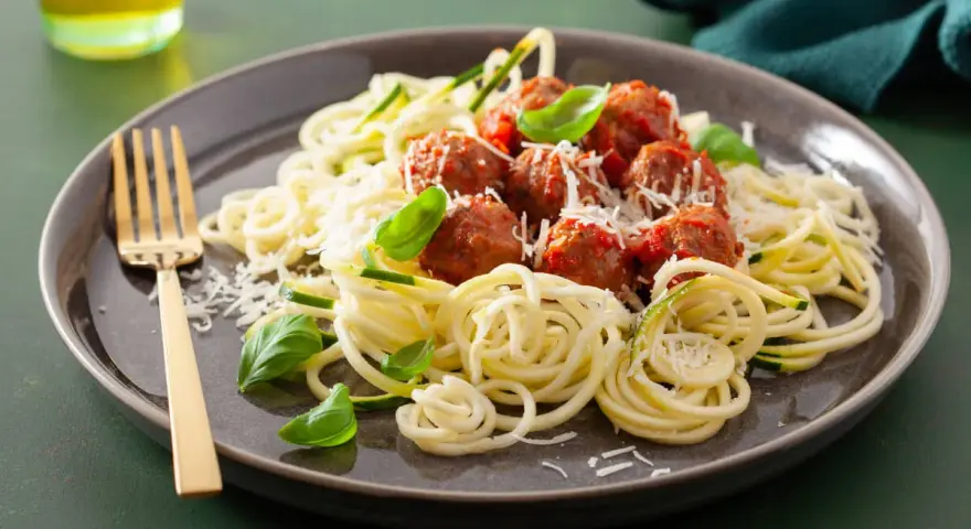 Pyszna keto kolacja: klopsiki w sosie pomidorowym na cukinii spaghetti, posypane parmezanem i listkami bazylii. Złoty widelec czeka.