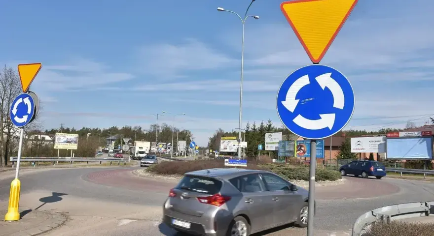 Rondo z tablicami "ustąp pierwszeństwa" i "ruch okrężny". Kierowca w szarym samochodzie może dostać mandat za wymuszenie pierwszeństwa.