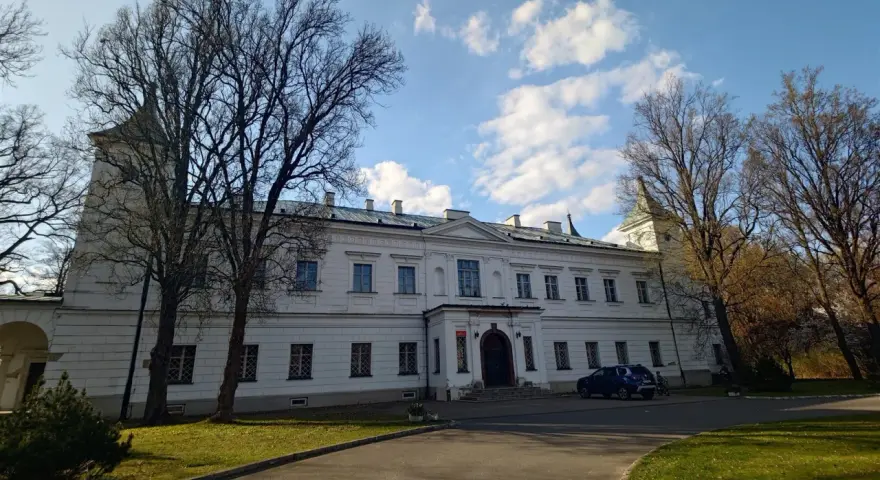 Pałac w Falentach, biała, okazała budowla z wieżyczką, otoczona drzewami o nagich gałęziach, pod błękitnym niebem z chmurami.