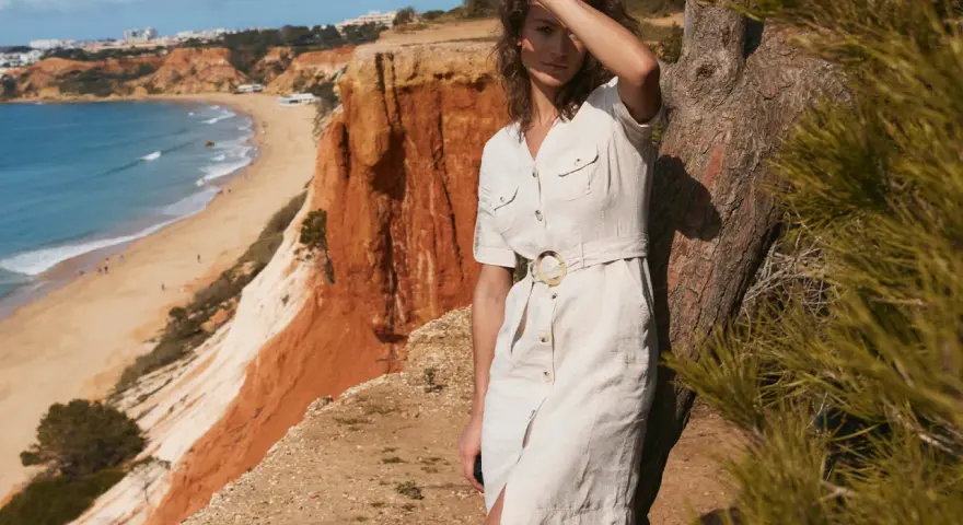 Kobieta w lnianej szmizjerce z paskiem stoi przy drzewie na klifie nad plażą.