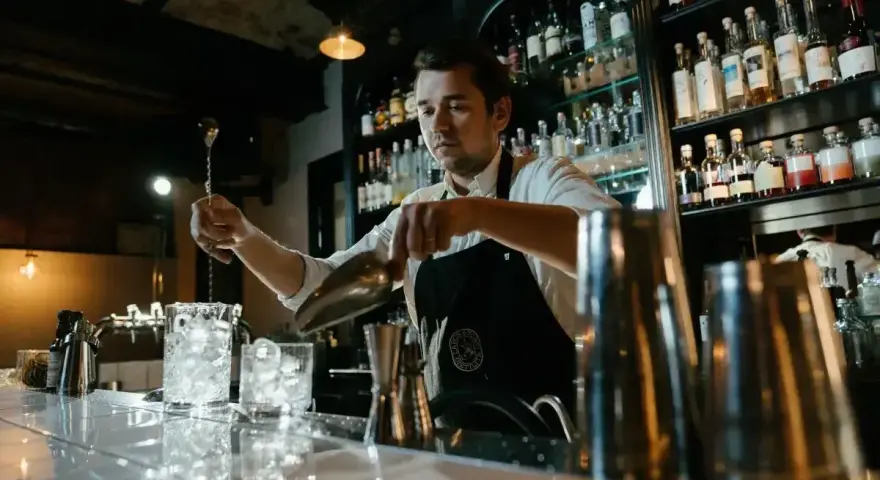 Barman przygotowuje drinki, pokazując, jak założyć drink bar. Na półkach stoją butelki alkoholi.