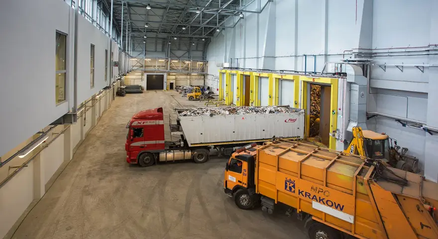Wielka hala przemysłowa, gdzie trwa termiczne przekształcanie odpadów. Ciężarówki z odpadami czekają na rozładunek, a koparka przygotowuje materiał do przetworzenia.