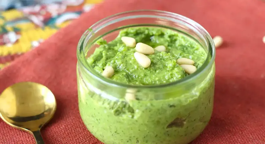 Frisches Rucola Pesto in einem Glas, garniert mit Pinienkernen. Daneben eine goldene Löffel.