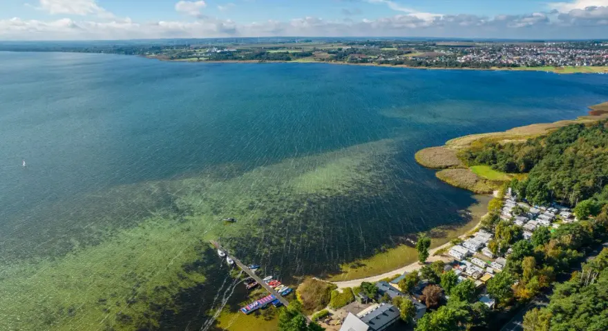 Malowniczy cypel na Helu z kempingiem, pomostem i kolorowymi łódkami. W oddali widać miasto i farmę wiatraków.