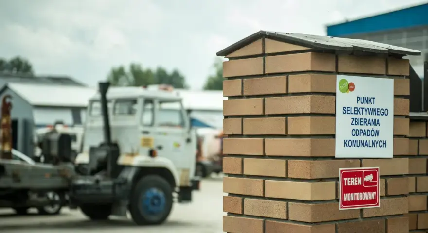 Tablica informuje, co to jest pszok – punkt selektywnego zbierania odpadów komunalnych. W tle widać ciężarówki.