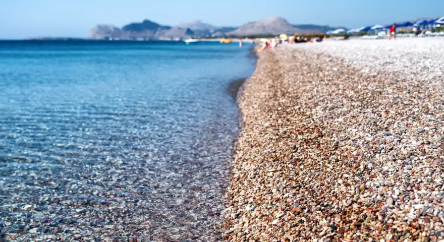 Rodos: turkusowe morze obmywa kamienistą plażę, w tle góry i plażowicze. Idealne miejsce na wakacje.