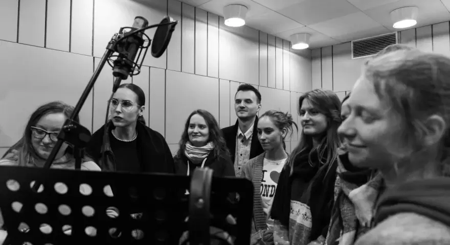 Grupa osób w studiu nagraniowym, przygotowująca podkładanie głosu do bajek. W tle mikrofon studyjny.