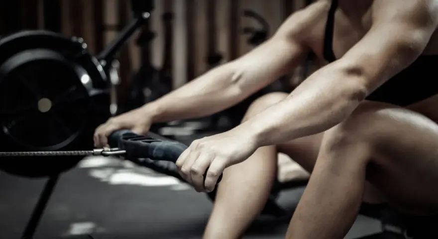 Une personne concentrée sur son entraînement, en plein effort pour faire du rameur. Ses bras sont tendus, ses mains agrippent la poignée.
