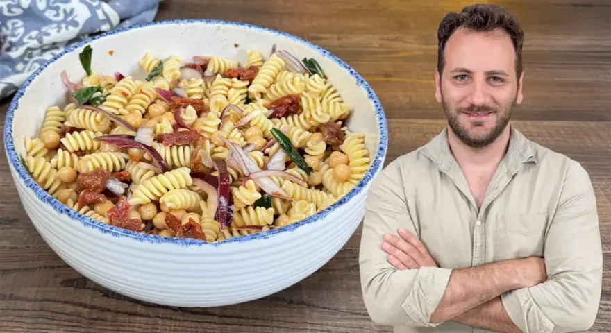 Un uomo sorride accanto a una ciotola di pasta fredda con ceci, pomodori secchi e cipolla rossa.