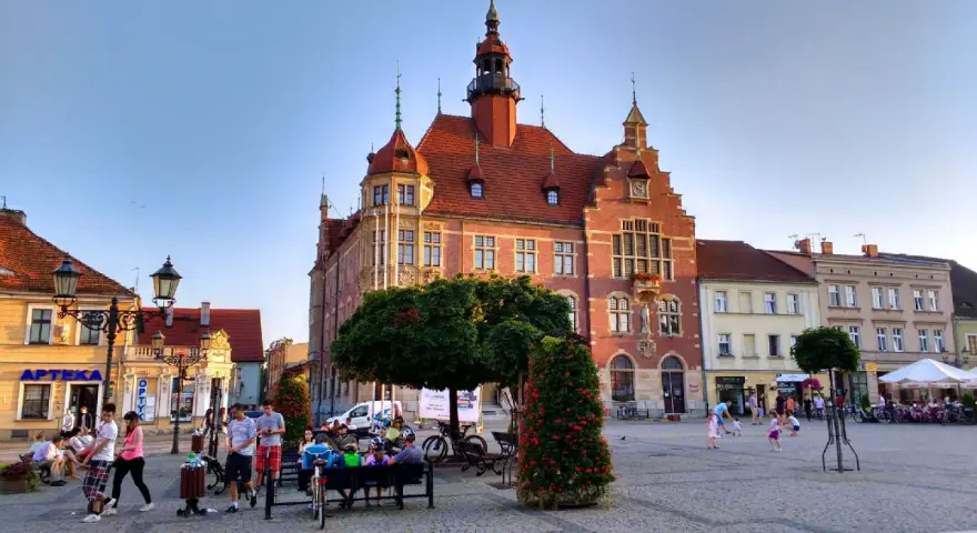 Rynek w Tarnowskich Górach tętni życiem. Ludzie spacerują, siedzą na ławkach, a dzieci bawią się na bruku. W tle widać zabytkowy ratusz.