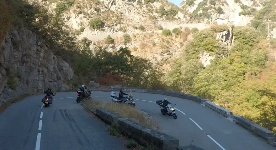 Motociclisti affrontano le curve del Col de Turini, immersi in un paesaggio montano roccioso e verdeggiante.