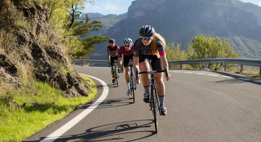 Three cyclists race up a mountain road. The lead cyclist, a woman, is what is a sportive person, focused and determined.