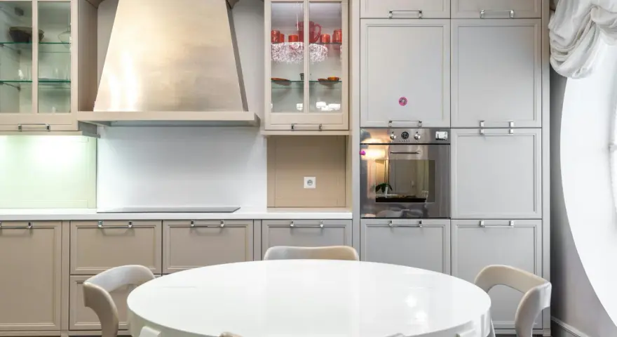 Cocina moderna con mesa redonda blanca y sillas. Gabinetes grises y beige, campana extractora de acero inoxidable. Ideal para ver catálogos de cocinas y precios.