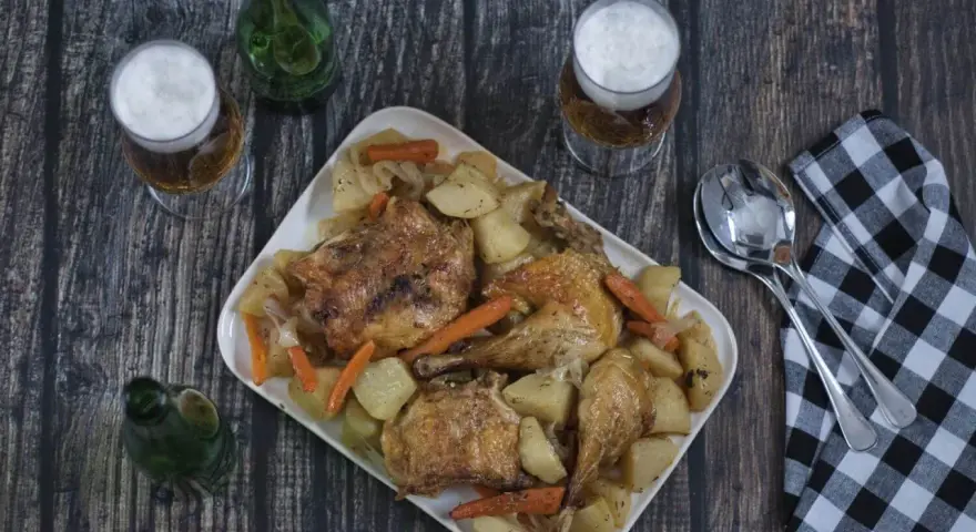 Pollo asado con patatas y zanahorias en una bandeja blanca, con cerveza y servilleta a cuadros.