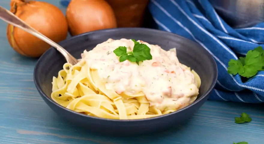 En skål med tagliatelle täckt av krämig ost och skinksås, garnerad med persilja. Två gula lökar syns i bakgrunden.