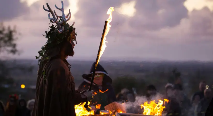En person med hjorthorn och lövverk håller en fackla vid en eld. En mystisk stämning under halloween i Irland.