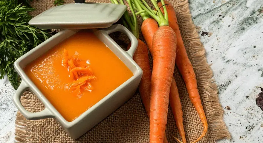 Un tazón de cremoso puré de zanahoria, adornado con hebras de zanahoria rallada, junto a zanahorias frescas y verdes.