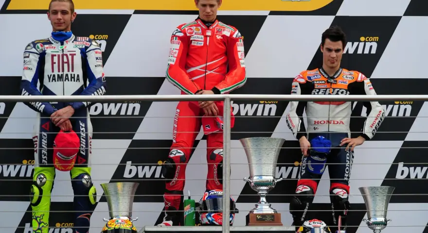 Podio con tre motociclisti famosi in tuta: Rossi, Stoner e Lorenzo. Vittoria celebrata con trofei.