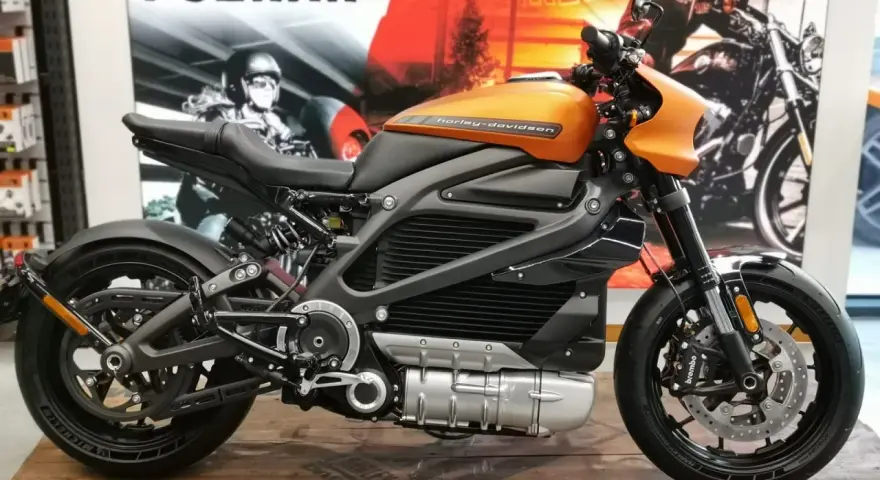 Pomarańczowy harley elektryczny z czarnymi akcentami, prezentowany na drewnianej podłodze, z plakatem Harley-Davidson w tle.