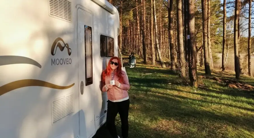 Kobieta z kubkiem kawy opiera się o kampera na leśnej polanie. Wspaniała przygoda w **Finlandii kamperem**!