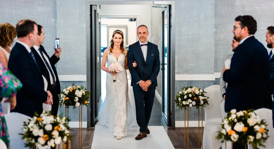 Panna młoda w sukni ślubnej idzie z ojcem do ołtarza, obok bukiety kwiatów.