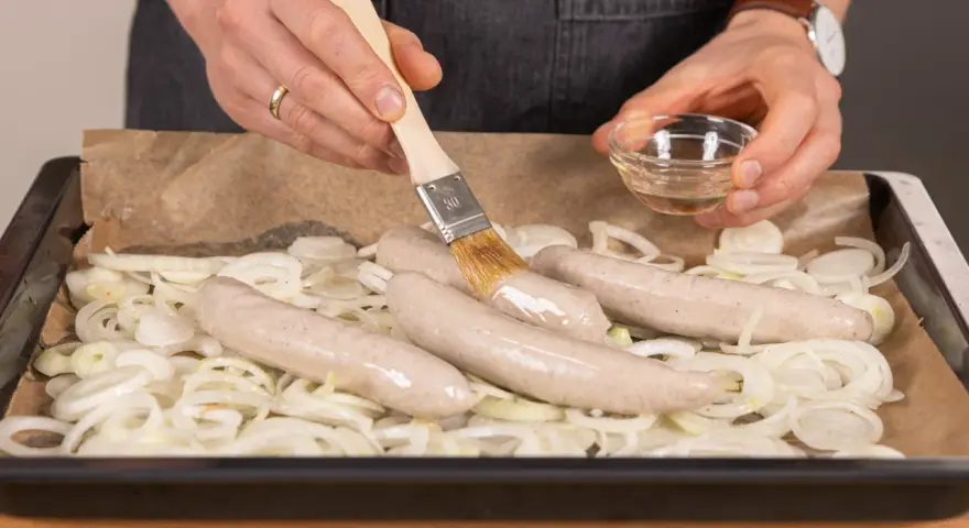 Bratwurst im Backofen: Zwiebelringe auf Backpapier, darauf vier rohe Bratwürste. Mit einem Pinsel wird Öl aufgetragen.