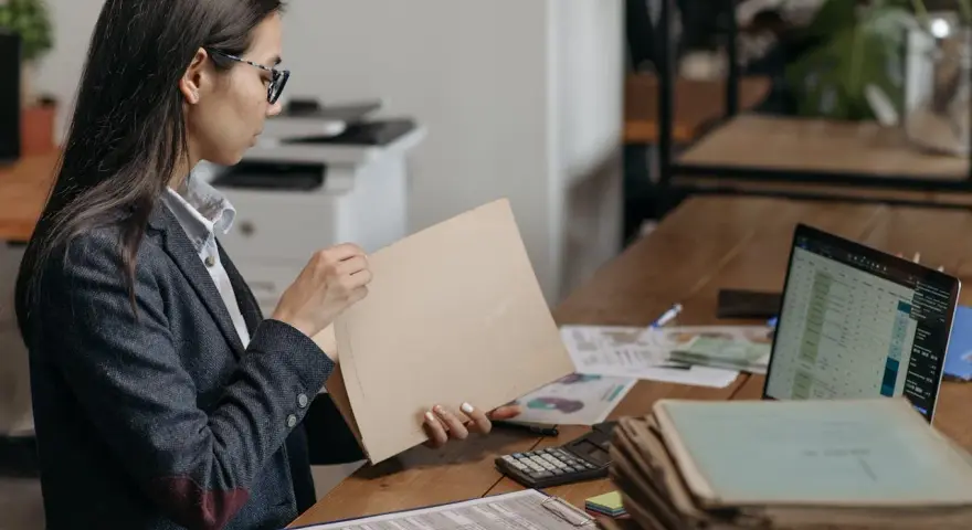 Kobieta w okularach przegląda dokumenty, szukając tytułu prawnego do lokalu. Na biurku leżą też laptop, kalkulator i sterty papierów.