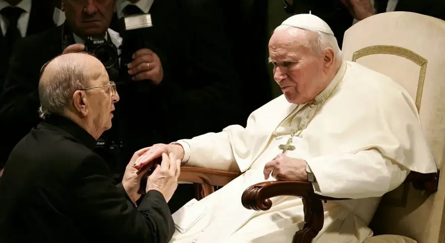 Papież Jan Paweł II w białej szacie, klęczący przed nim mężczyzna w czerni. W tle widać innych duchownych i dziennikarzy, być może z zakonu **jezuitów**.