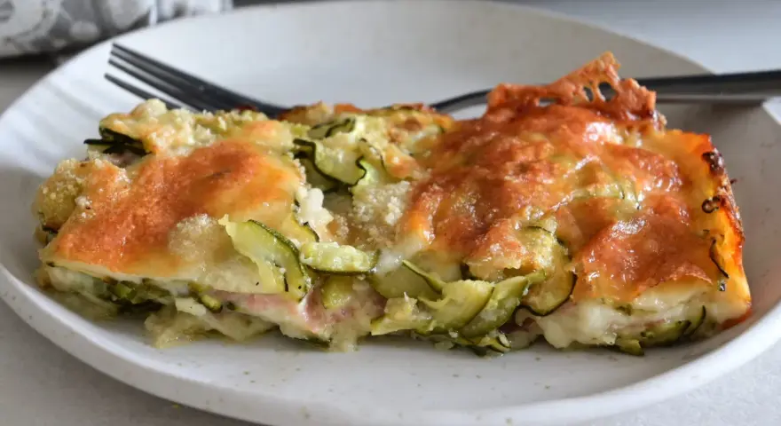 Una fetta di sformato di zucchine dorato e filante, con formaggio fuso e zucchine a rondelle, servito su un piatto bianco.