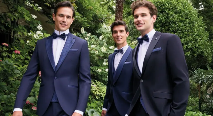 Tre eleganti sposo e testimoni in abiti scuri, sorridono in un giardino lussureggiante.