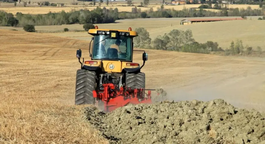 Trattore giallo in azione per l'aratura del terreno, sollevando terra e polvere in un paesaggio rurale collinare.