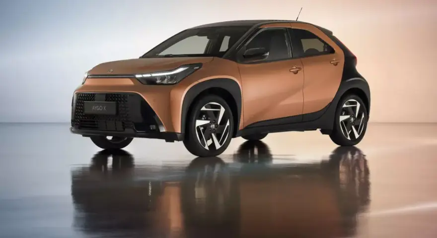 Brązowy Toyota Aygo X z czarnym dachem i felgami na błyszczącym podłożu.