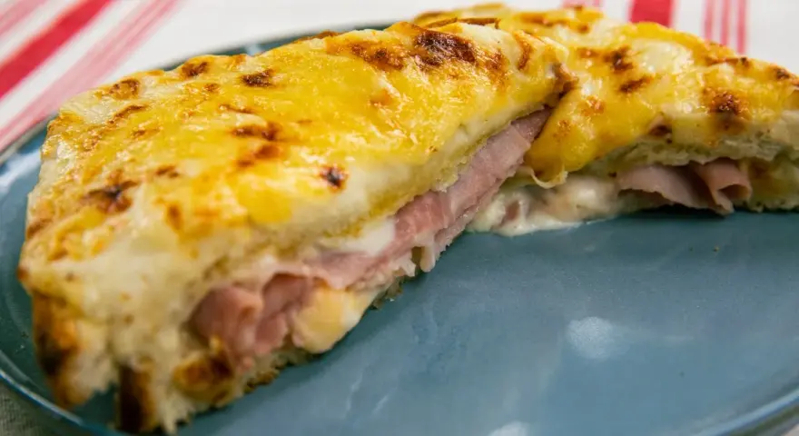 Croque Monsieur gratinado con queso derretido y jamón, servido en plato azul.