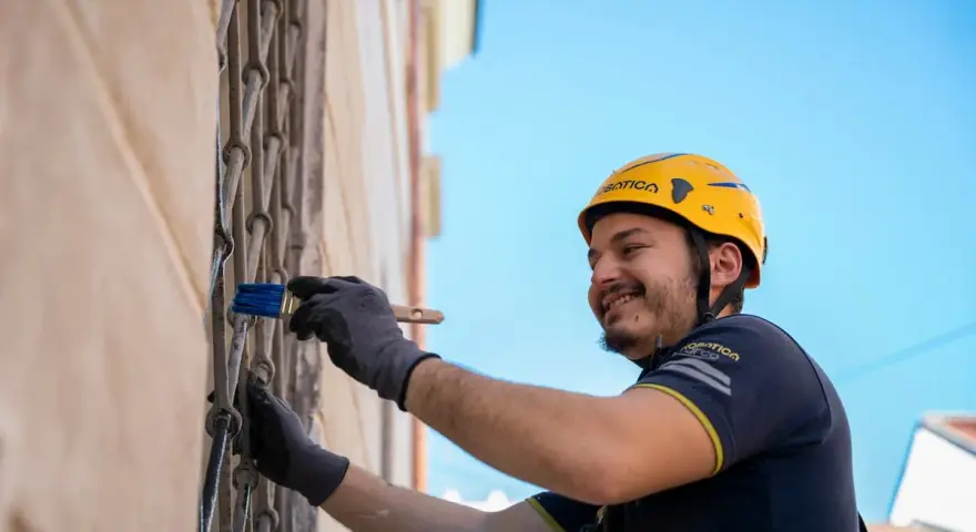 Uomo con casco giallo dipinge una grata metallica con un pennello blu. Inquadratura dal basso, cielo azzurro sullo sfondo.