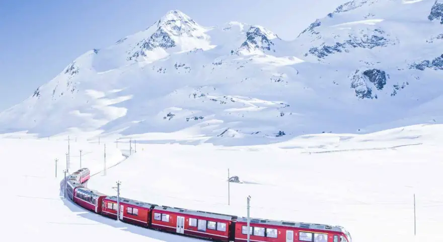 A bright red St. Moritz train winds through a pristine, snow-covered mountain landscape under a clear blue sky.