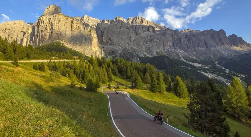 Motociclisti sfrecciano su una strada tortuosa verso il Passo Furcia, circondati da verdi pendii e imponenti montagne rocciose sotto un cielo azzurro.