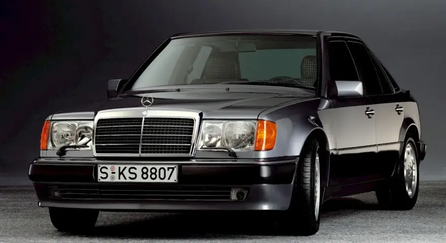 Klasyczny Mercedes W124 w ciemnym kolorze, z charakterystyczną atrapą chłodnicy i reflektorami. Ten model słynie z niezawodnych silników.