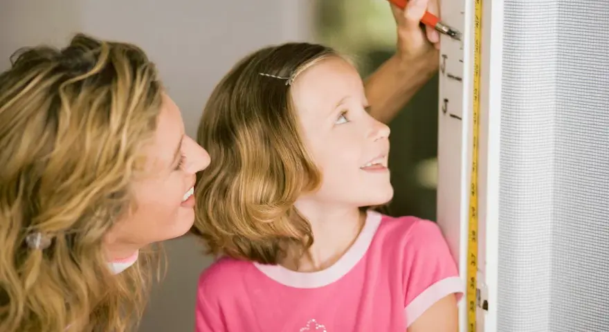 Mama mierzy córkę, która z zaciekawieniem patrzy na miarkę. Czyżby zastanawiała się, dlaczego dziecko nie rośnie tak szybko, jak by chciała?