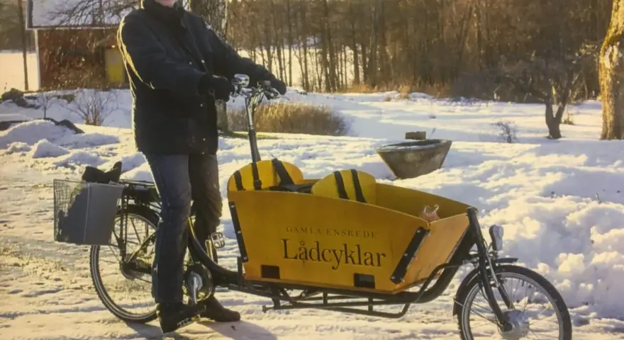 Man med mössa och jacka cyklar på vintern med en gul lastcykel i snöig miljö.