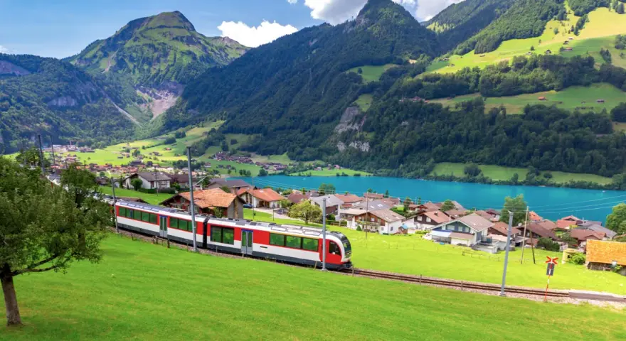 A red and white train travels through a picturesque Swiss village nestled beside a turquoise lake, with lush green mountains in the background. This is the scenic route from Lucerne to Lugano.