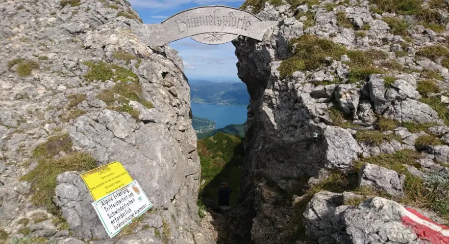 Himmelspforte am Schafberg: Ein Felsentor mit Holzinschrift, das den Blick auf einen See freigibt. Ein Wanderer steht im Durchgang.