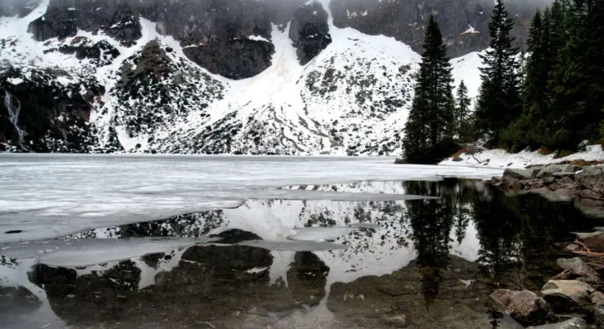 Morskie Oko zimą, zamarznięte, z odbiciem ośnieżonych gór i ciemnych drzew w przejrzystej wodzie.