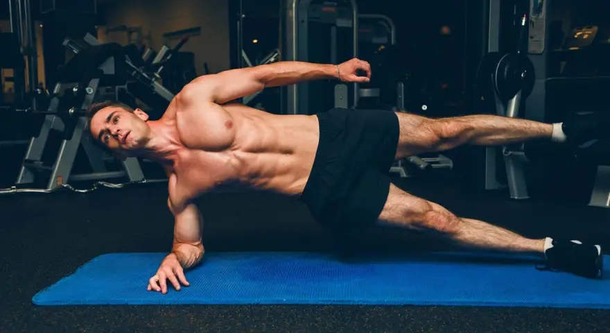 Un homme musclé réalise un gainage planche sur un tapis bleu dans une salle de sport.