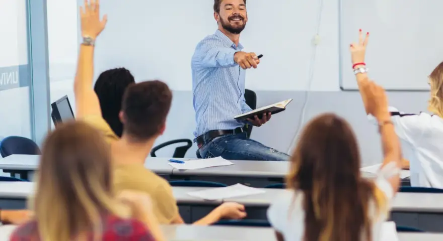 Uśmiechnięty wykładowca z książką w ręku wskazuje na studentów, którzy podnoszą ręce. Czy przed lecz stawiamy przecinek?